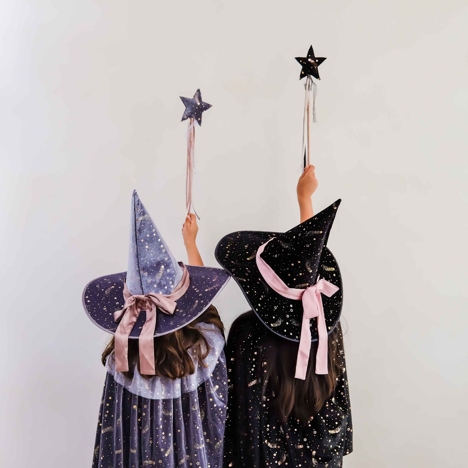Two children from behind wearing witch hats and dresses, holding star-shaped wand toppers against a plain light background.